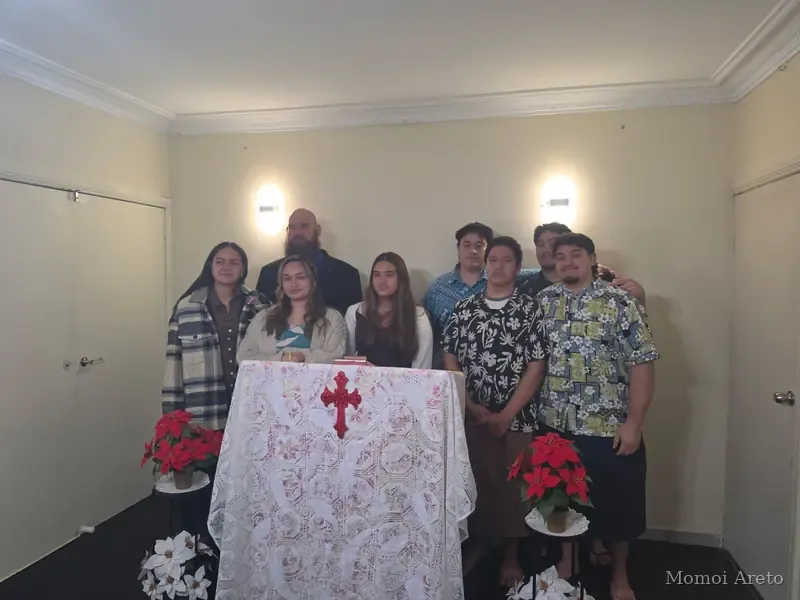 People standing in a plain cream-walled room behind a table covered with a white lace cloth with a red cross on it, with red poinsettia plants on either side.