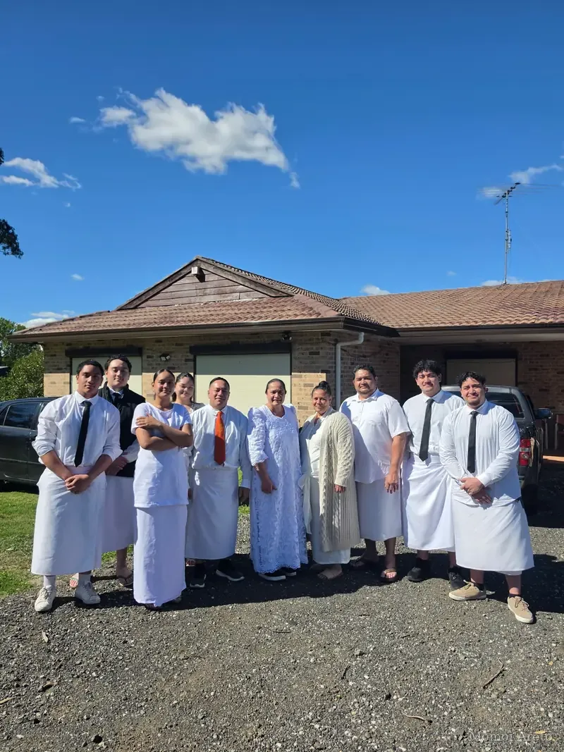 A line of people dressed in white, the men in white shirts and the women in long white dresses, standing on a gravel driveway in front of a single-storey brick house with a tiled roof, under a clear blue sky.